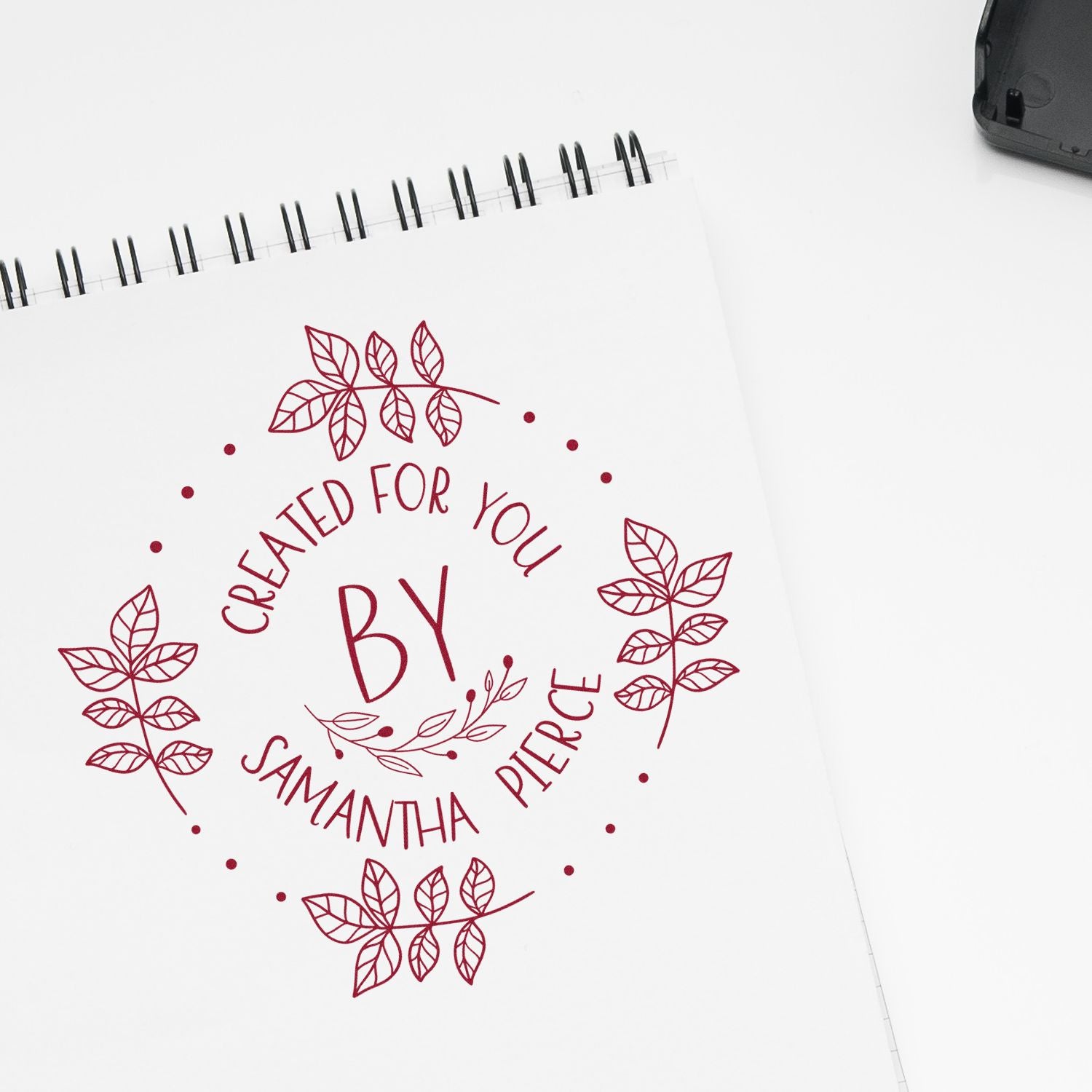 Maroon custom stamp and seal design with leafy wreath, reading Created for You by Samantha Pierce, on white spiral notebook.