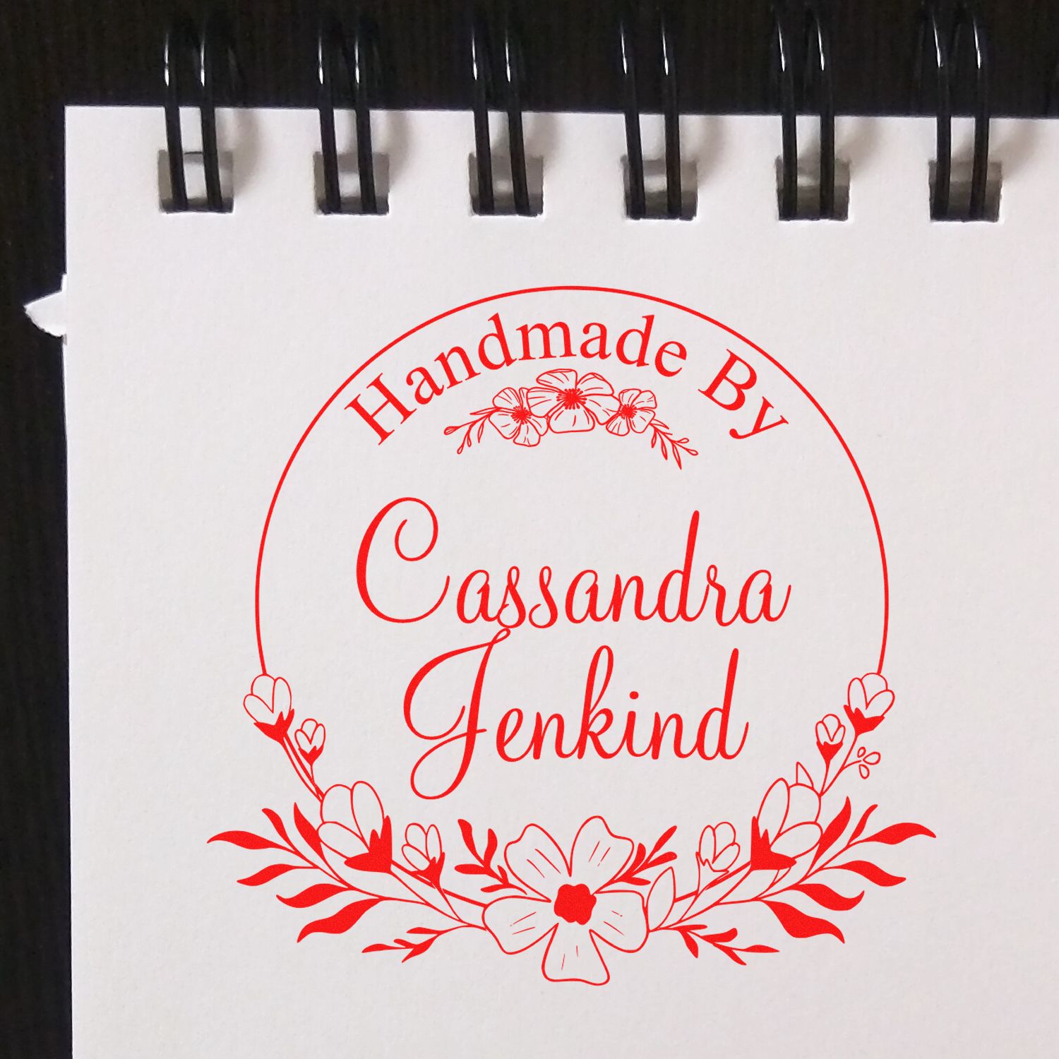 Red handmade stamp seal with floral wreath reading Handmade By Cassandra Jenkind on a white spiral notebook page