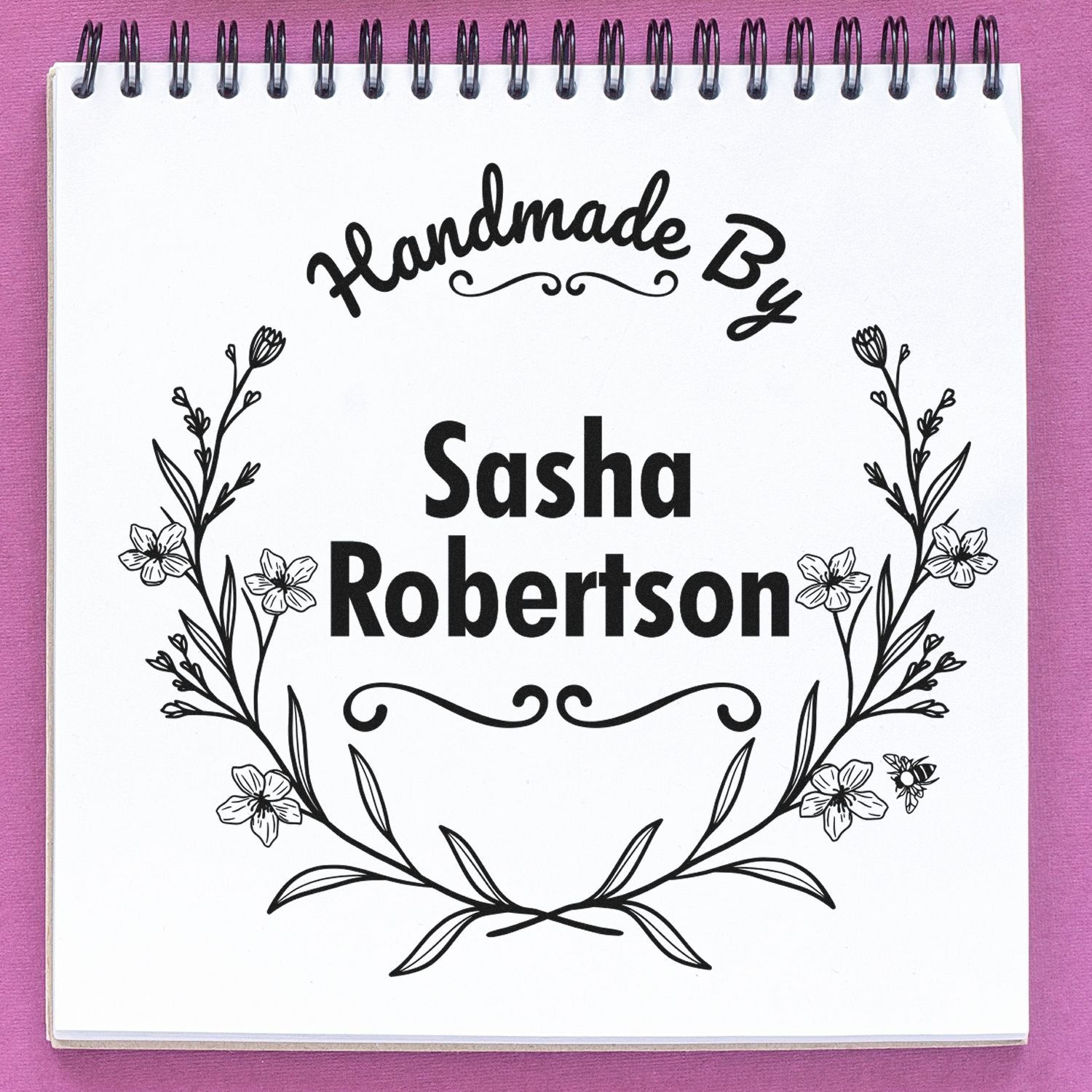 Personalized black handmade stamp seal with floral wreath and Sasha Robertson on white spiral notebook, pink background