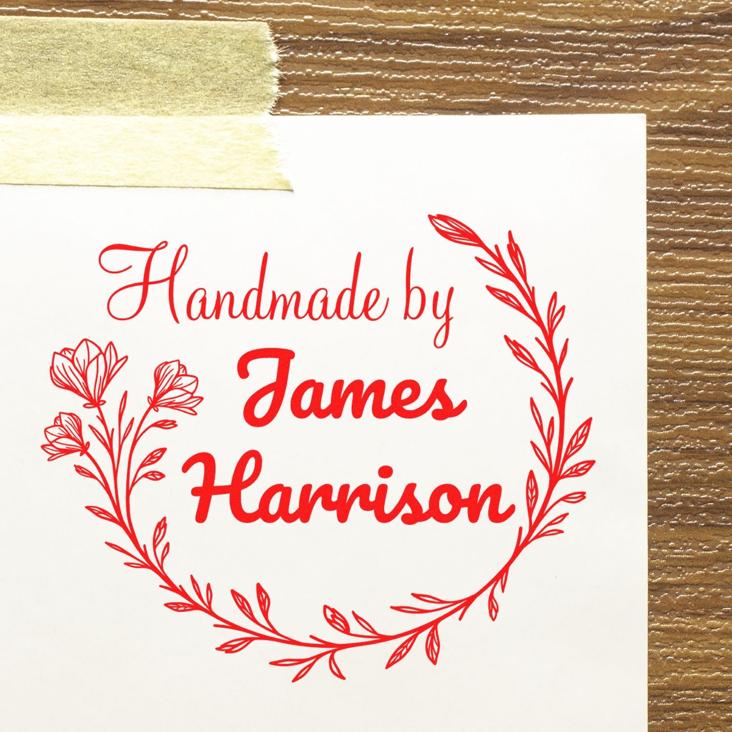 Red custom handmade stamp with floral wreath and James Harrison on white paper, taped to a brown textured surface.