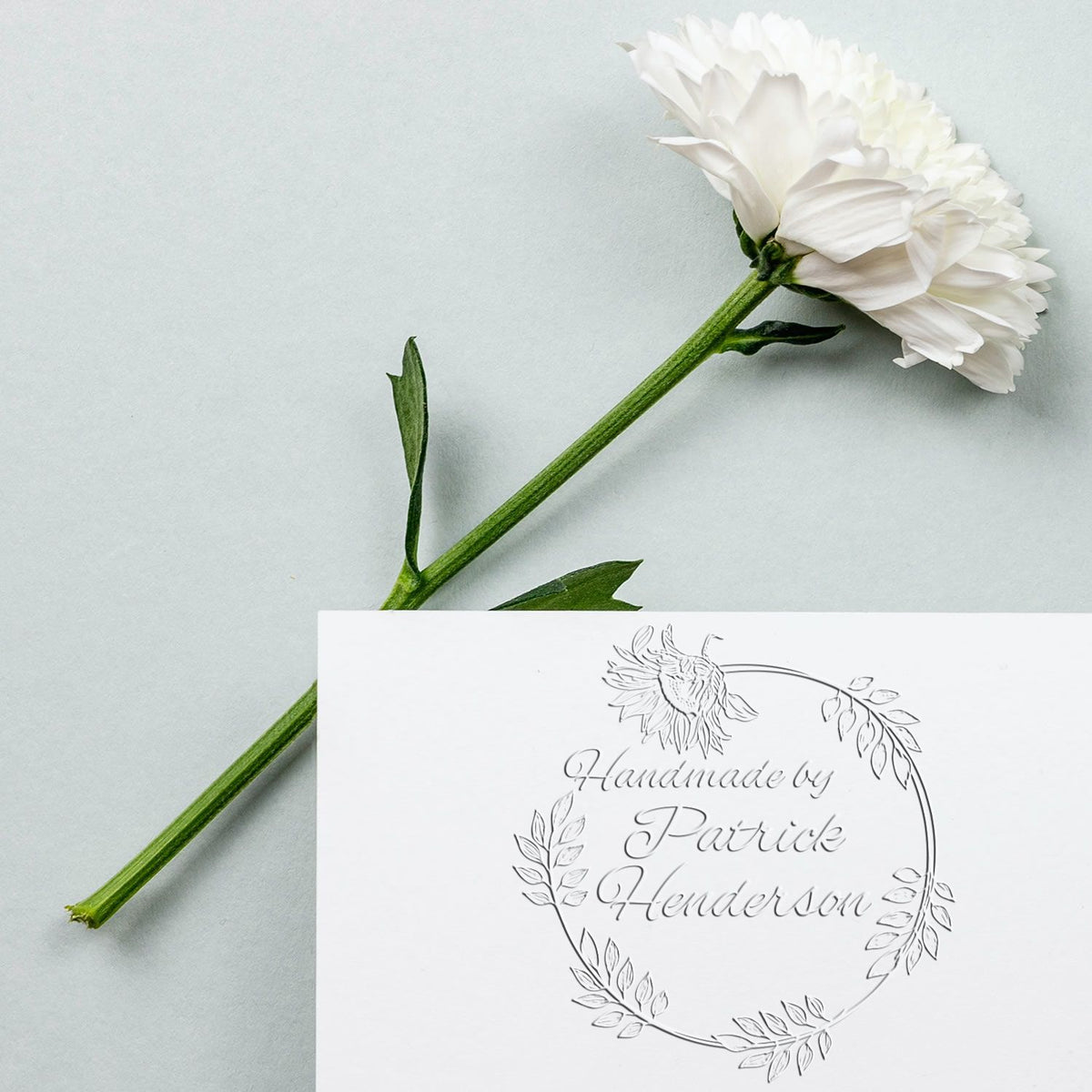 White floral custom stamp seal with green stem on pale blue background and Handmade by Patrick Henderson script.
