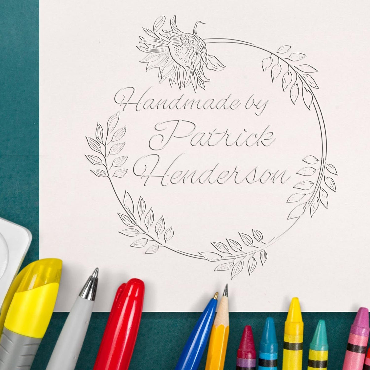 Custom stamp and seal design with floral wreath and embossed script on white paper, surrounded by colorful pens.
