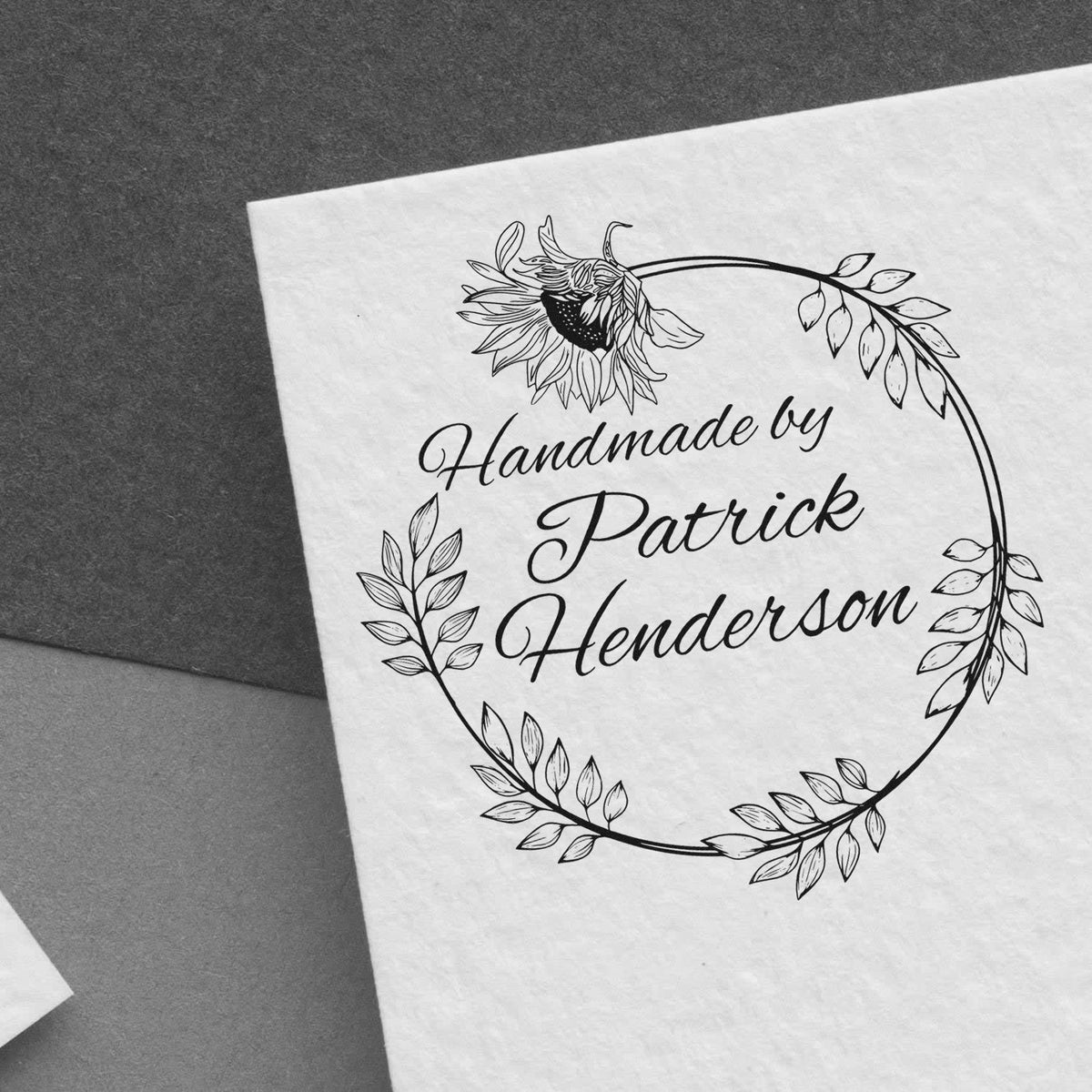 Black custom stamp seal with floral wreath, sunflower, and handwritten text Handmade by Patrick Henderson on white paper.