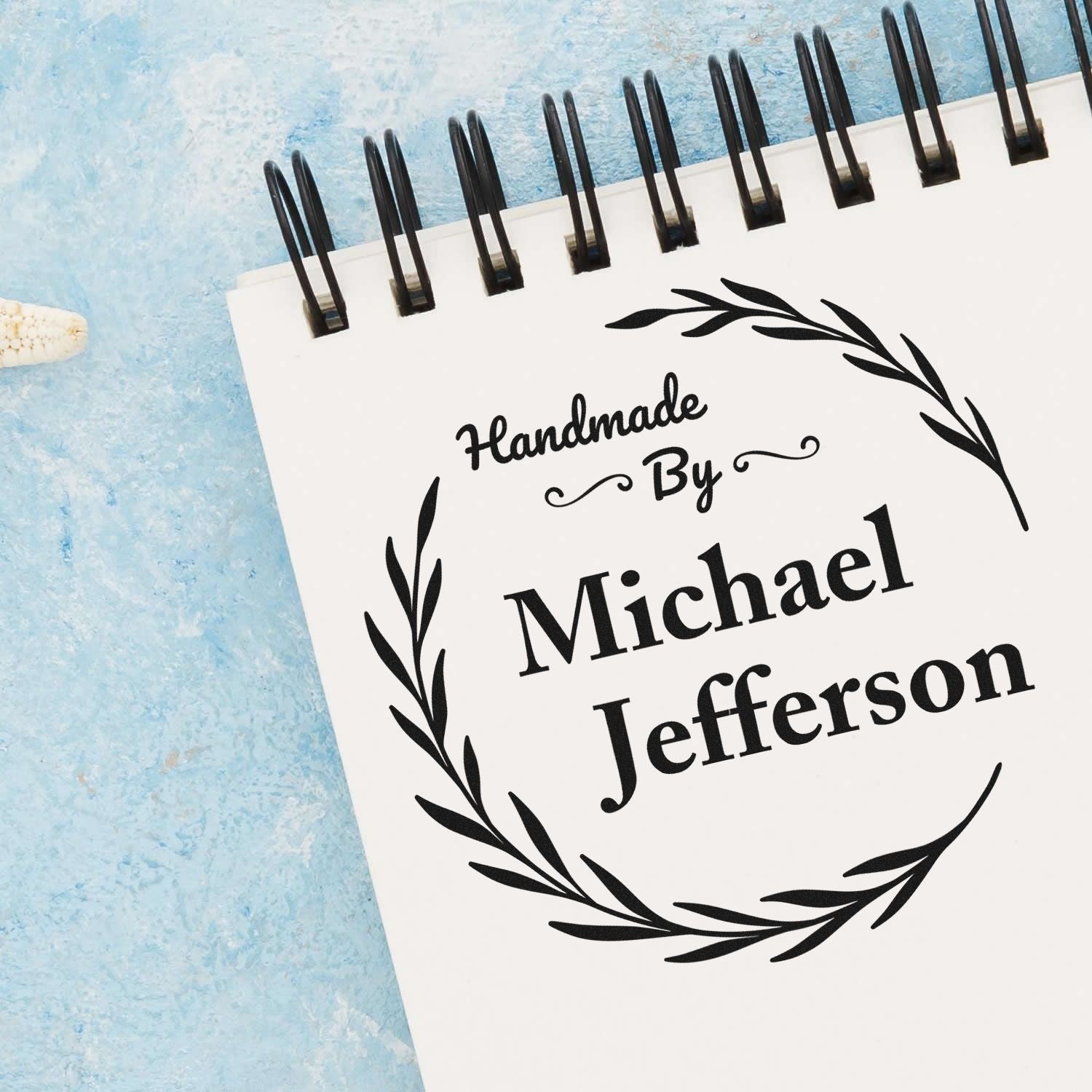 Custom black handmade stamp seal with laurel wreath and Michael Jefferson on a white notebook, against a blue textured background.