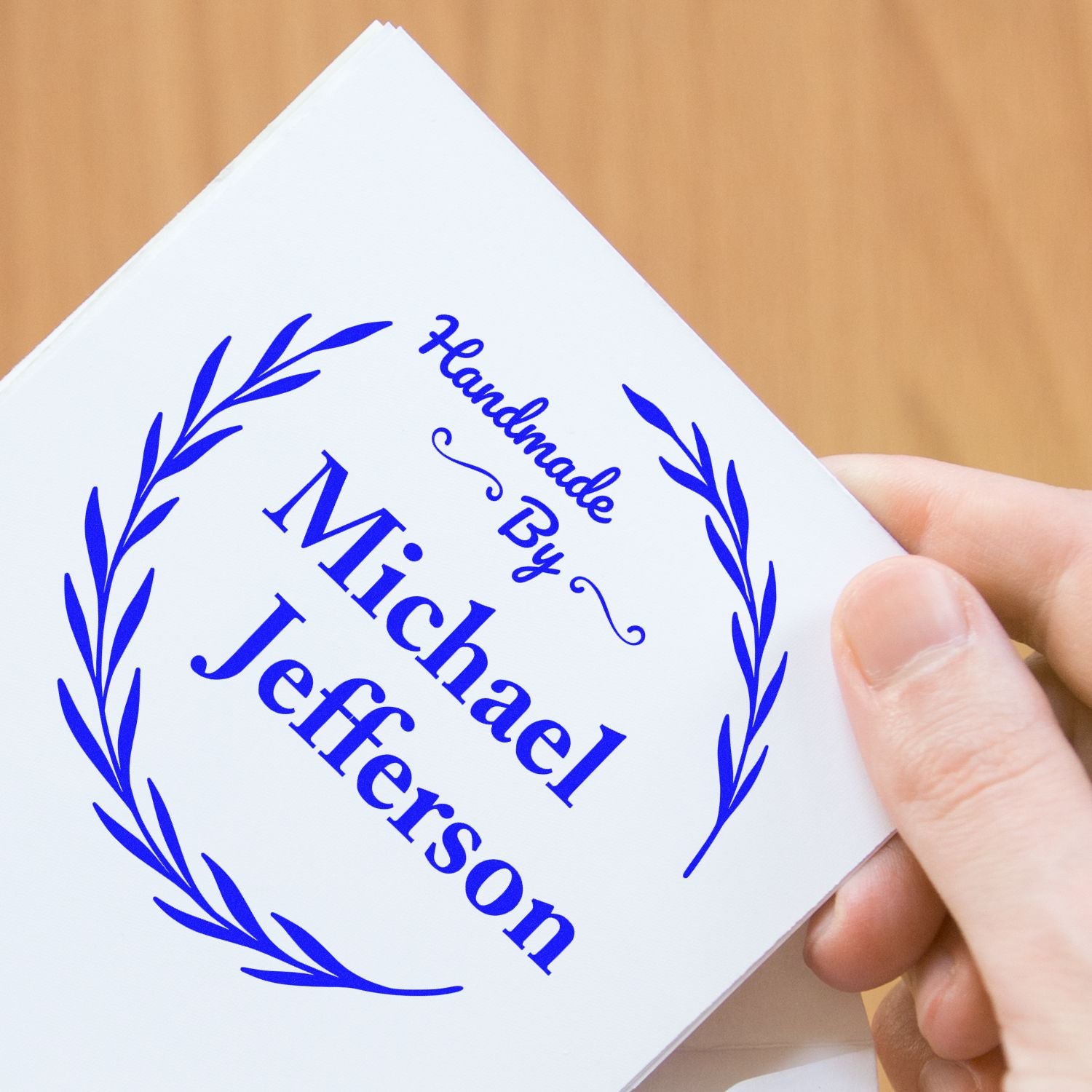 Blue custom handmade seal stamp with laurel wreath and ÂMichael Jefferson text on a white card, held in hand.