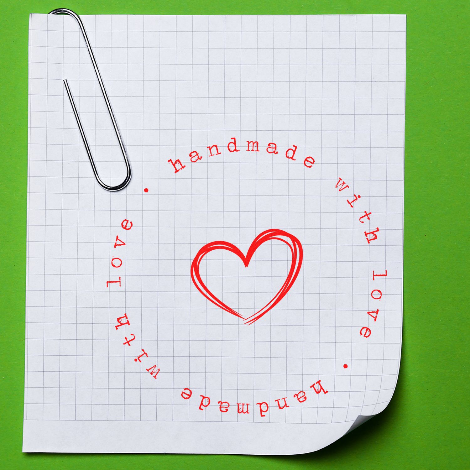 Red handmade-with-love heart stamp seal on gridded paper, with silver paper clip and green background