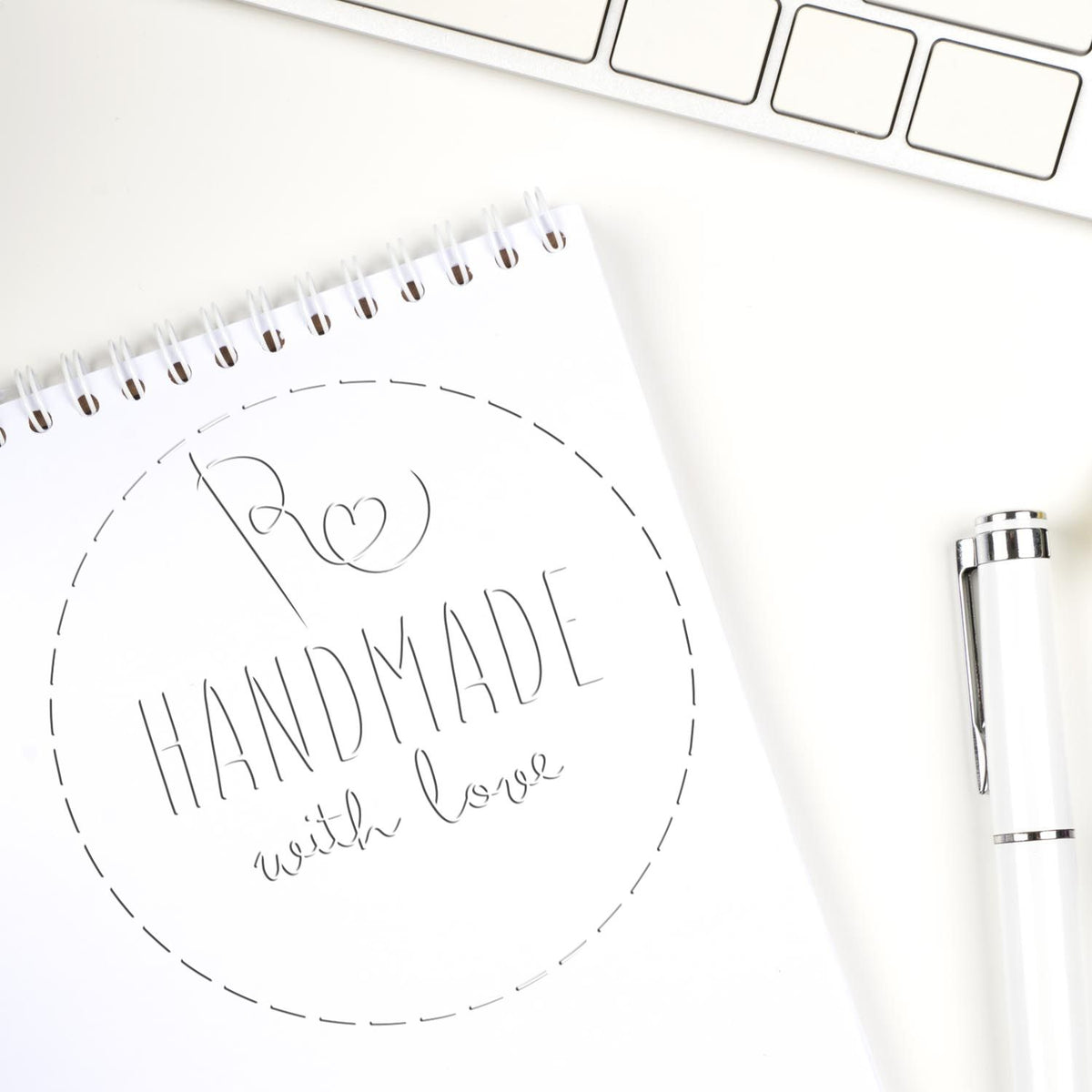 White custom stamp seal design on spiral notebook, with HANDMADE with love text and a white pen on a clean desk.