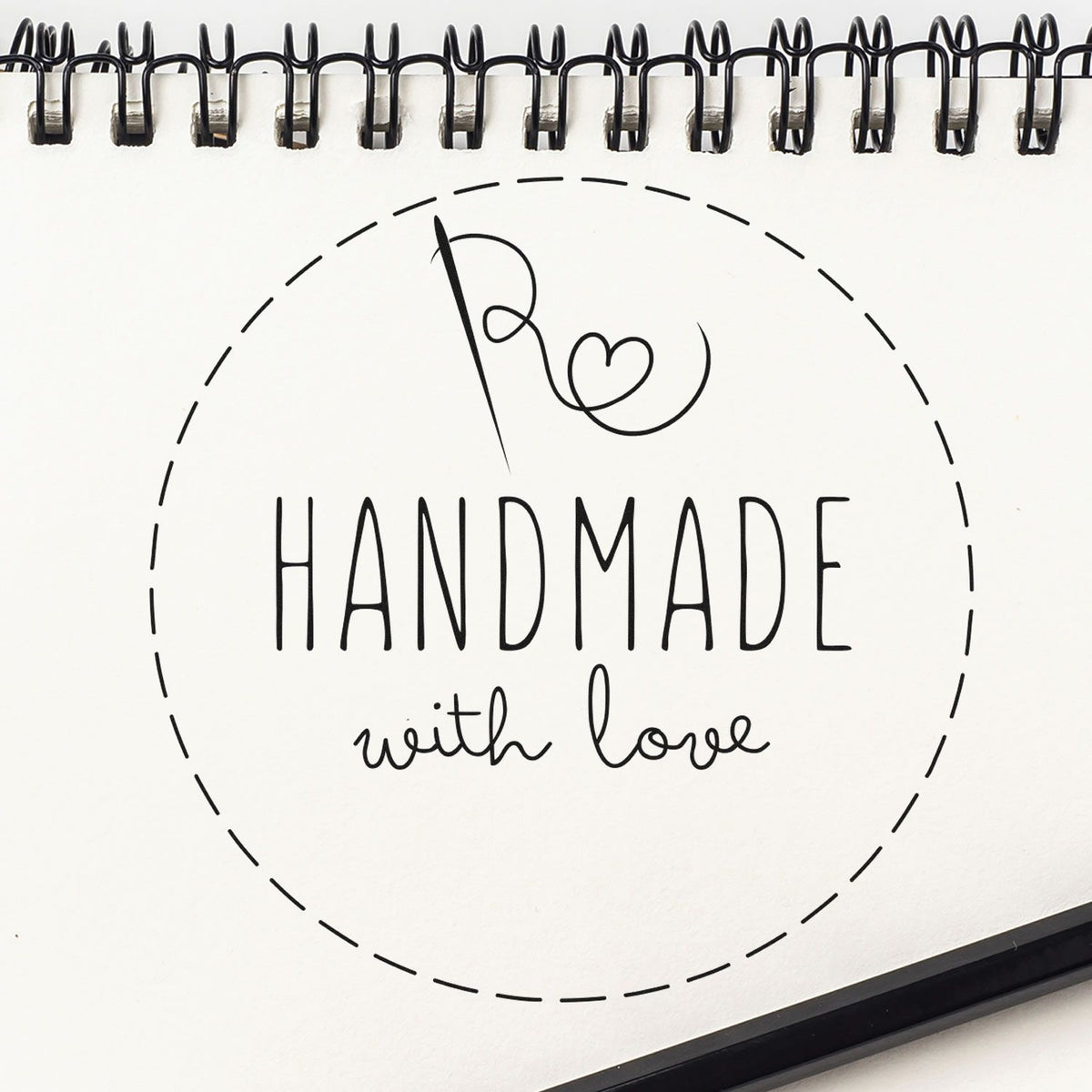 Black handmade with love stamp and seal design on white spiral notebook, with dashed circle and heart.