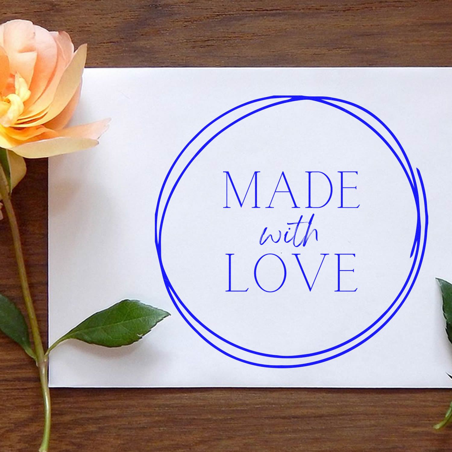 Blue round stamp and seal reading Made with Love on white paper, with peach flower and green leaves on a wood background.
