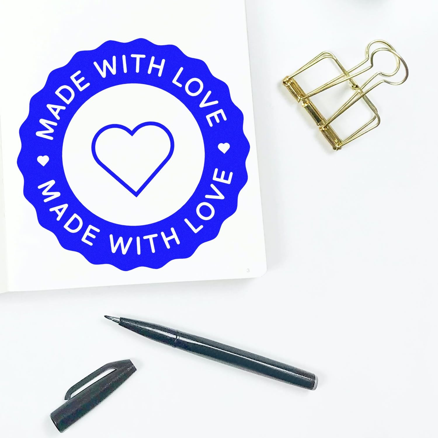 Blue Made with Love seal stamp with heart, beside gold binder clip and black pen on white background.