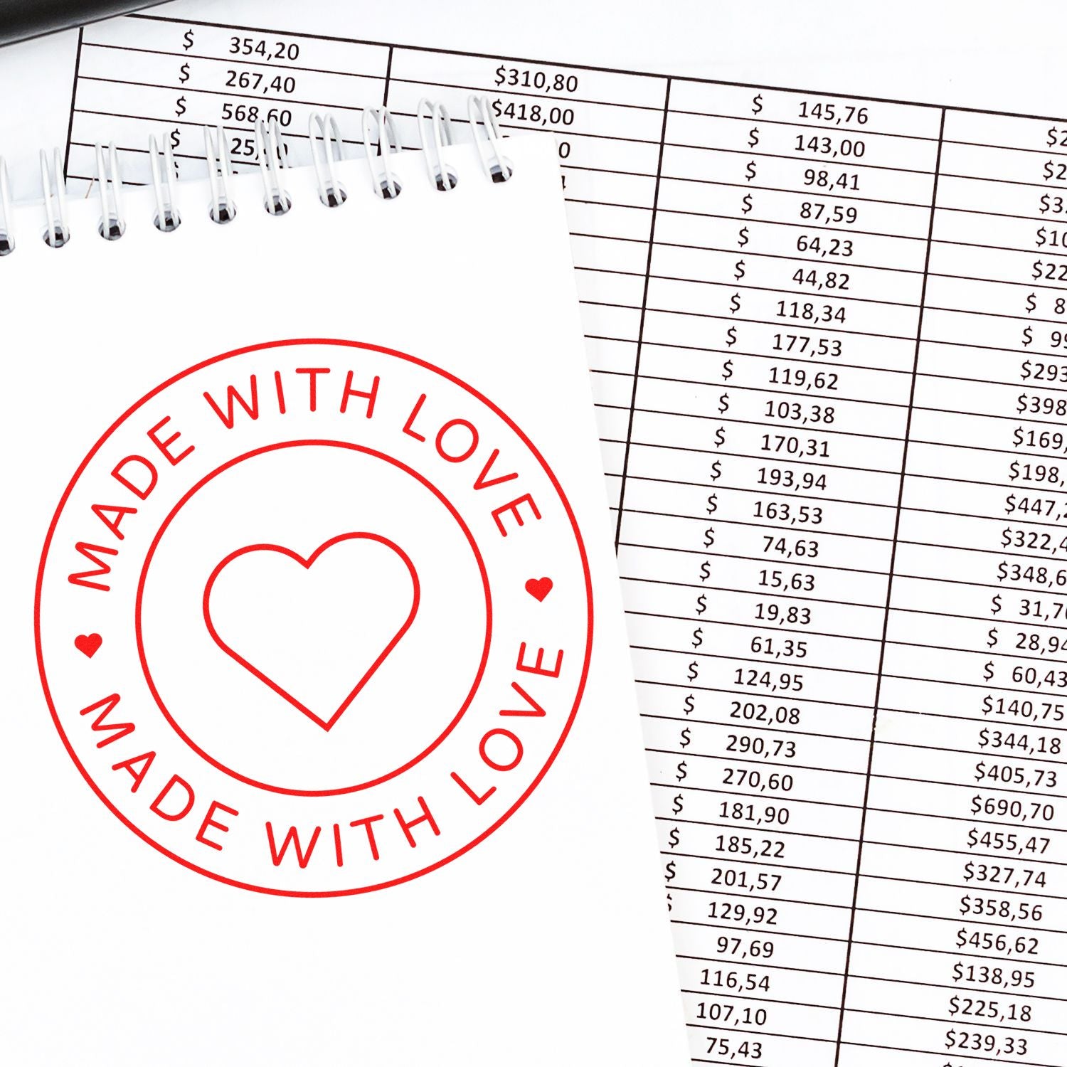 Red Made with Love round seal stamp with heart on a white spiral notepad over a financial spreadsheet.