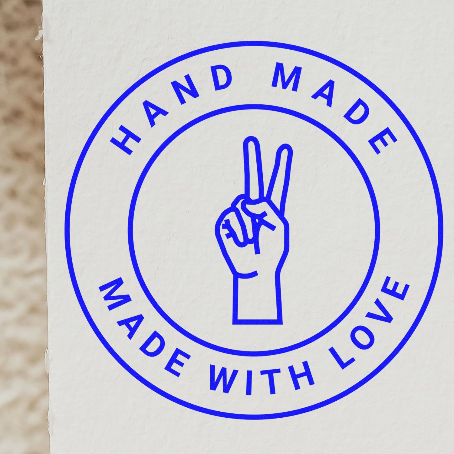 Blue handmade seal stamp with peace hand symbol and circular “Hand Made With Love” text on white paper.