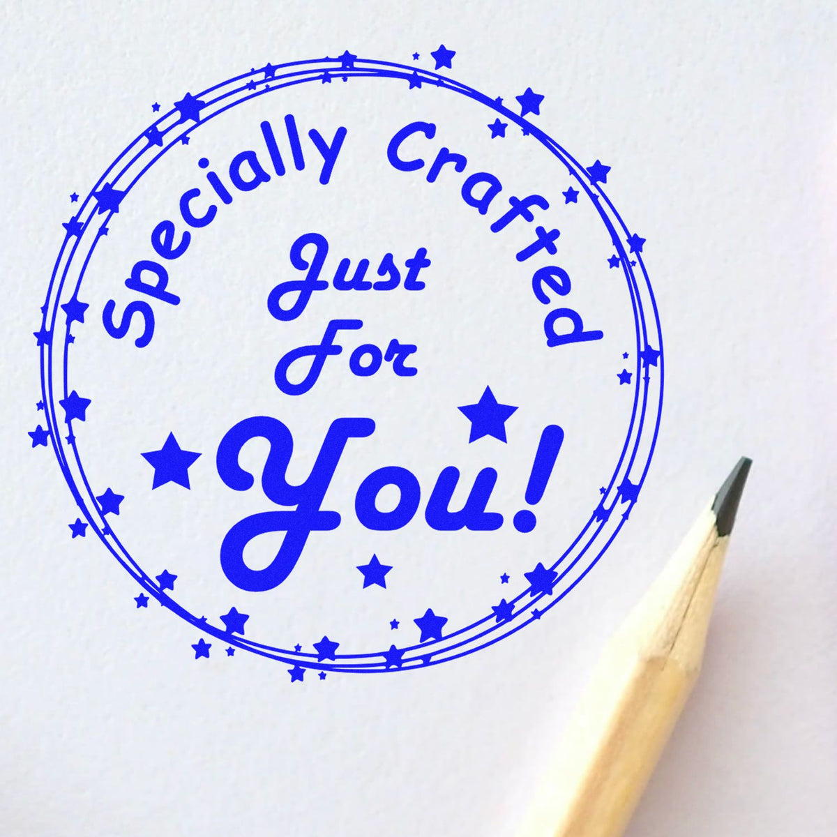 Blue circular stamp seal reading Specially Crafted Just For You! with stars on white paper, pencil tip at right.