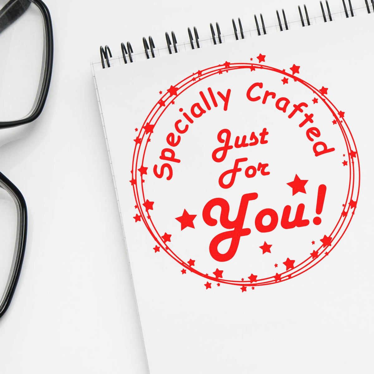 Red custom stamp seal reading Specially Crafted Just For You! on a white notebook, with black eyeglasses nearby.