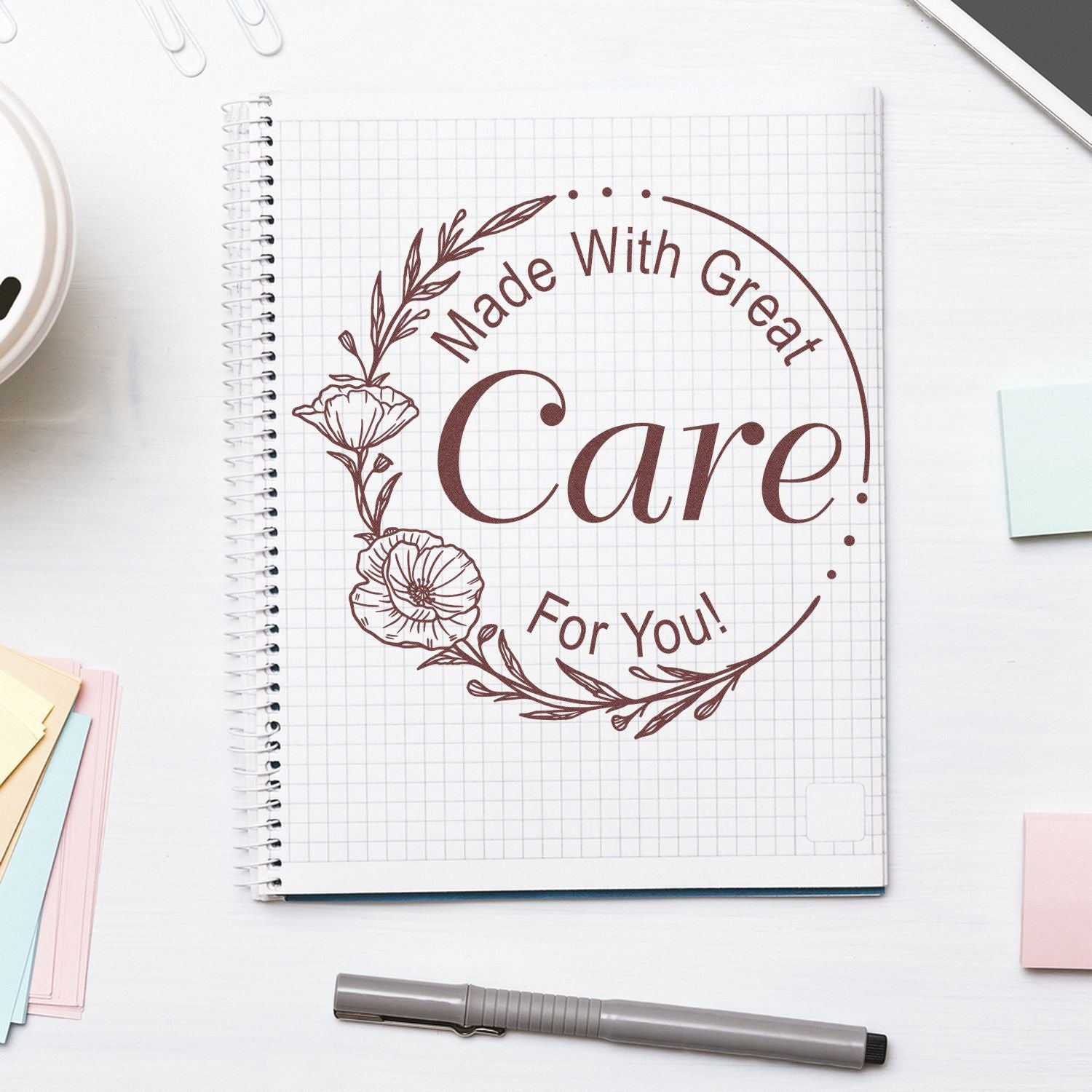 Brown floral stamp seal reading “Made With Great Care For You!” on a grid notebook, with pen and pastel paper.