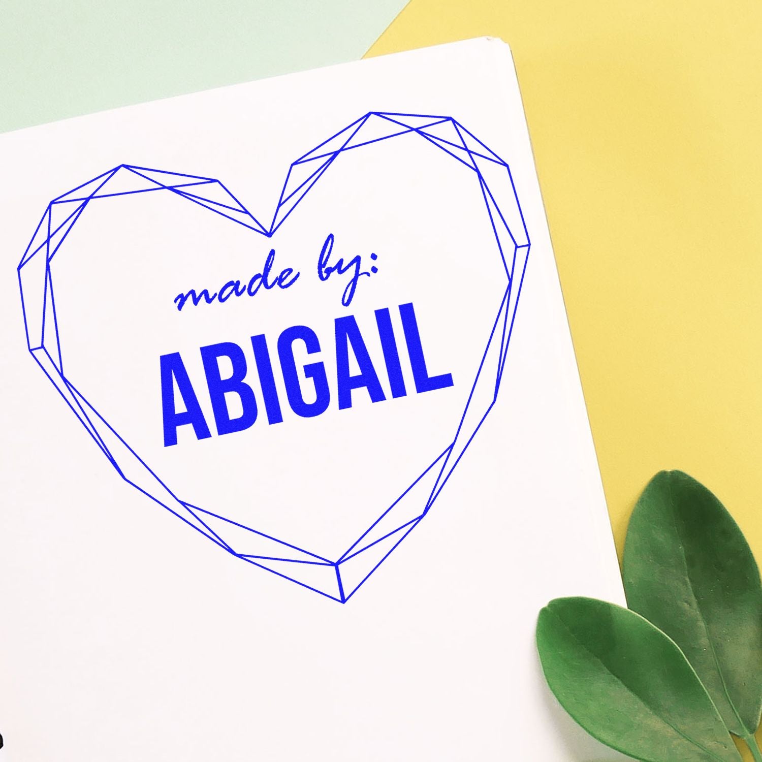 Blue geometric heart custom stamp and seal reading made by: ABIGAIL on white paper, with yellow background and green leaves.