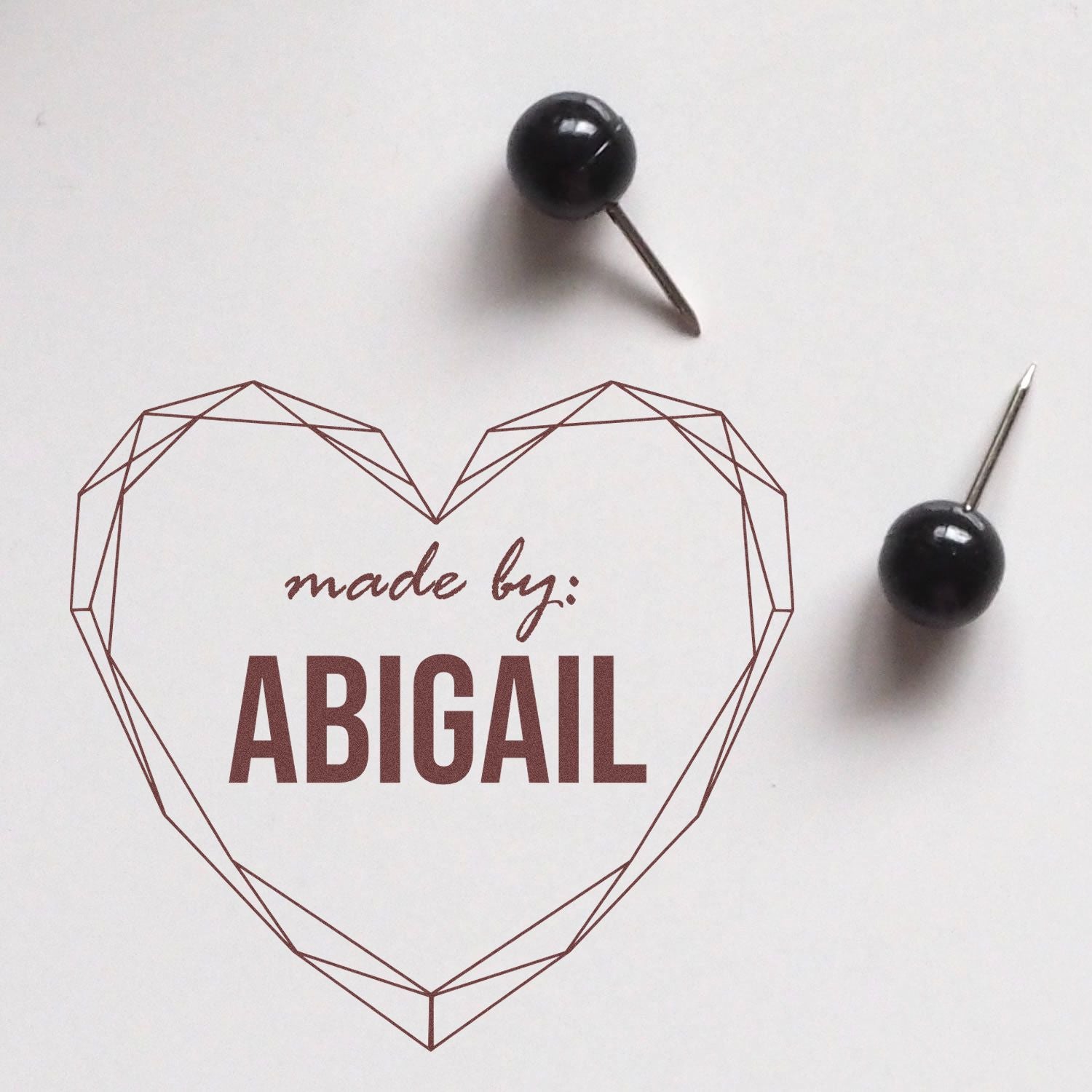 Burgundy heart-shaped stamp seal reading made by: ABIGAIL with two black pin-style seals on white.
