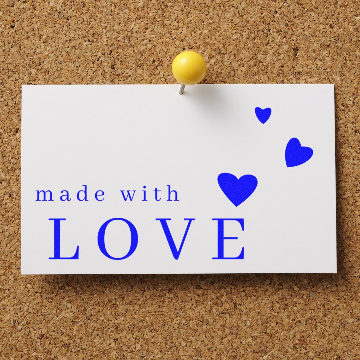 White stamp and seal card on corkboard with blue made with LOVE text and heart seals, pinned by a yellow pushpin.