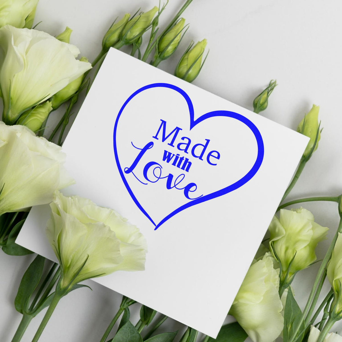 Blue heart-shaped “Made with Love” stamp seal on white card, framed by pale yellow-green flowers.