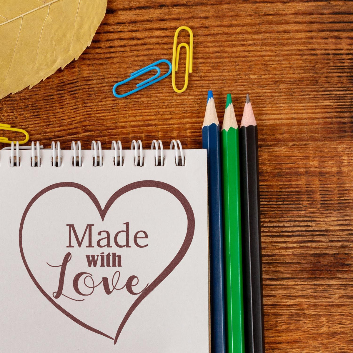 Heart-shaped brown stamp and seal reading “Made with Love” on notepad, with blue, green, and black pencils on wood.