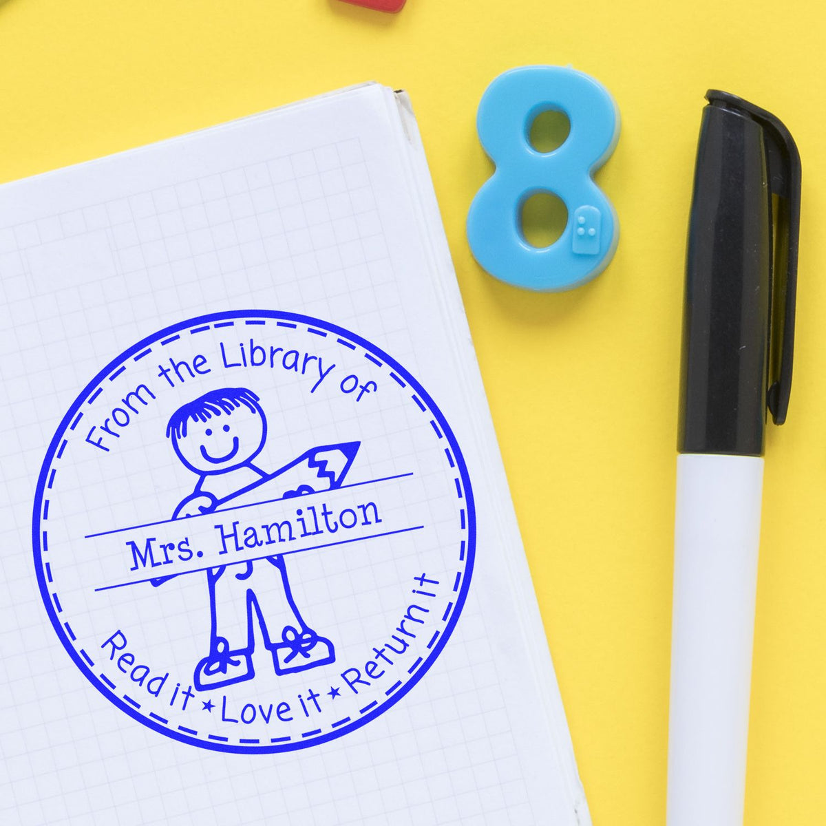 Blue custom library self-inking stamp and teal number 8 seal on notebook with black marker on yellow background.