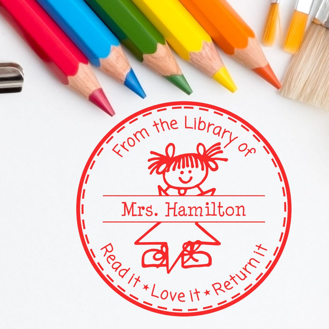 Red custom library stamp and return seal with a childrens drawing, surrounded by colorful pencils on a white background.