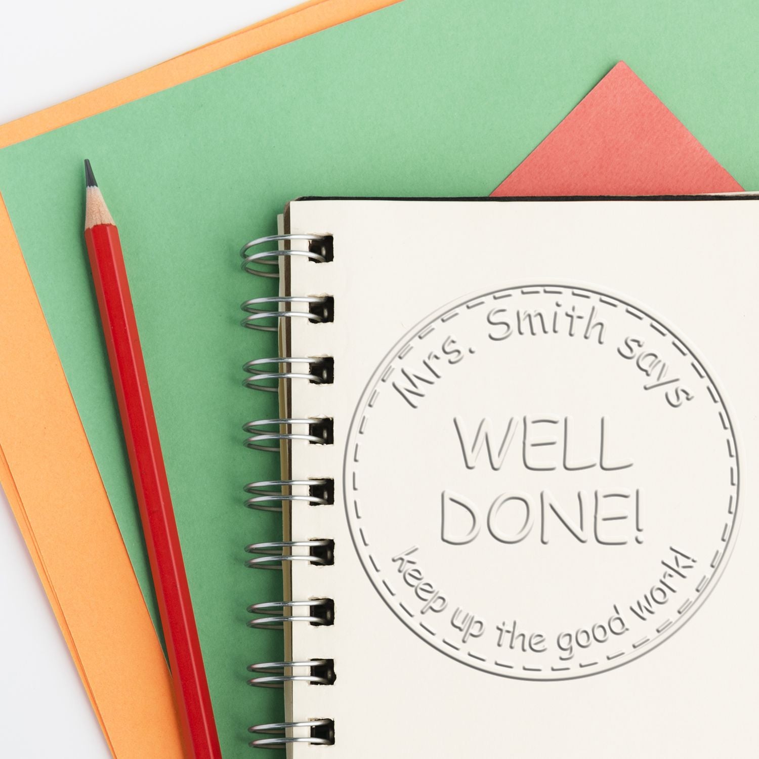 Circular rubber stamp seal on spiral notebook reading WELL DONE! beside a red pencil on green and orange paper.