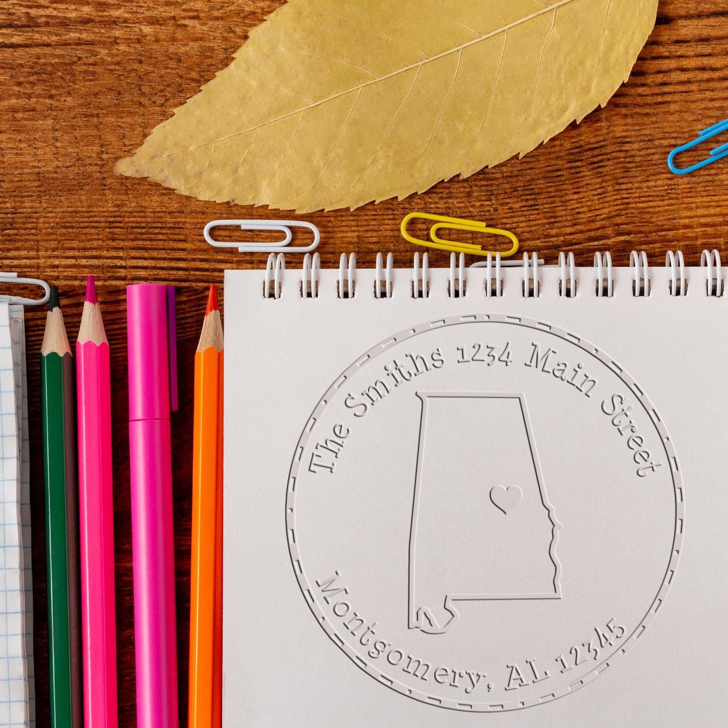 Custom embossed seal notebook with Alabama outline, colorful pencils, and tan leaf on rustic wood.