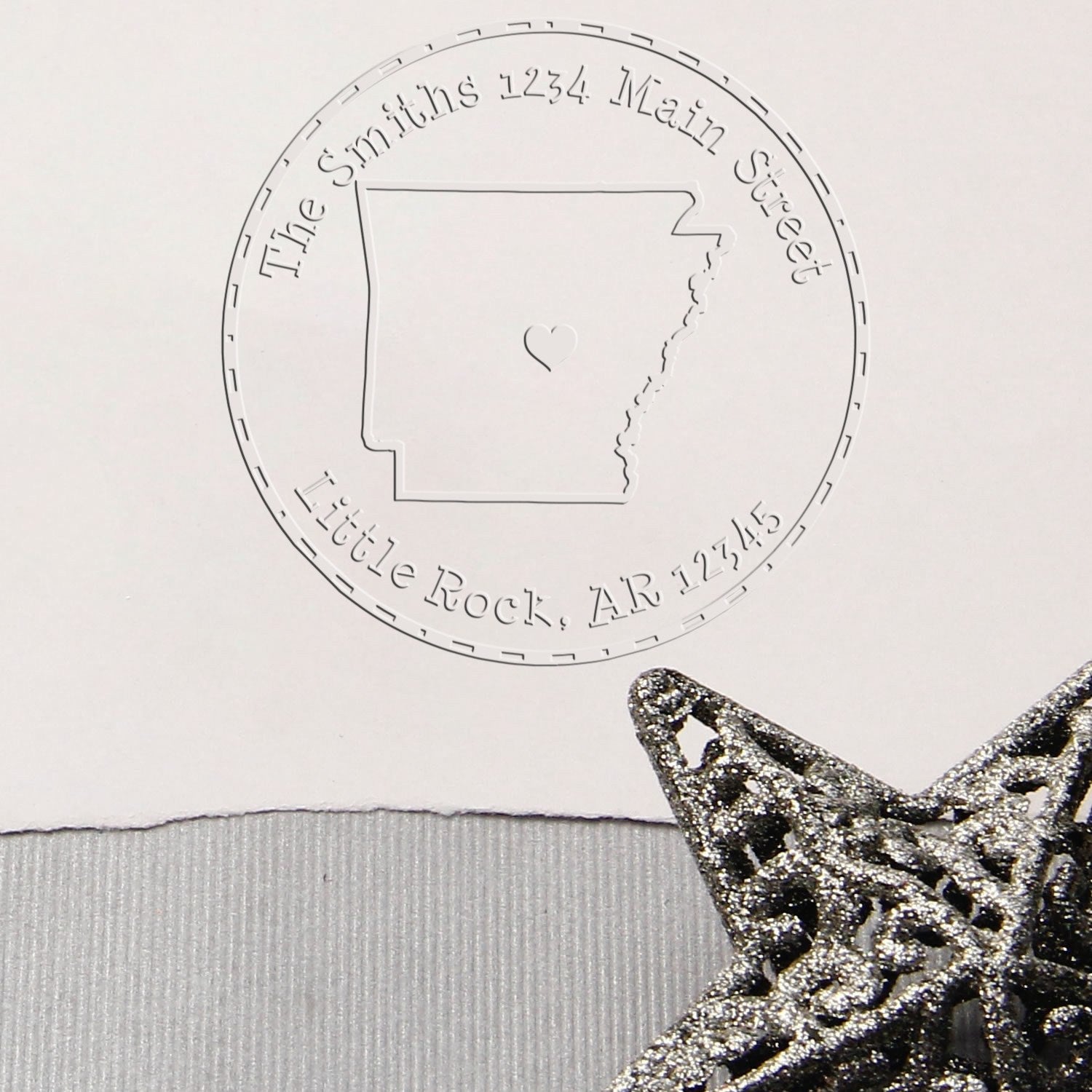 Embossed custom stamp and seal on white paper with Arkansas outline, circular address text, and glittery silver star.