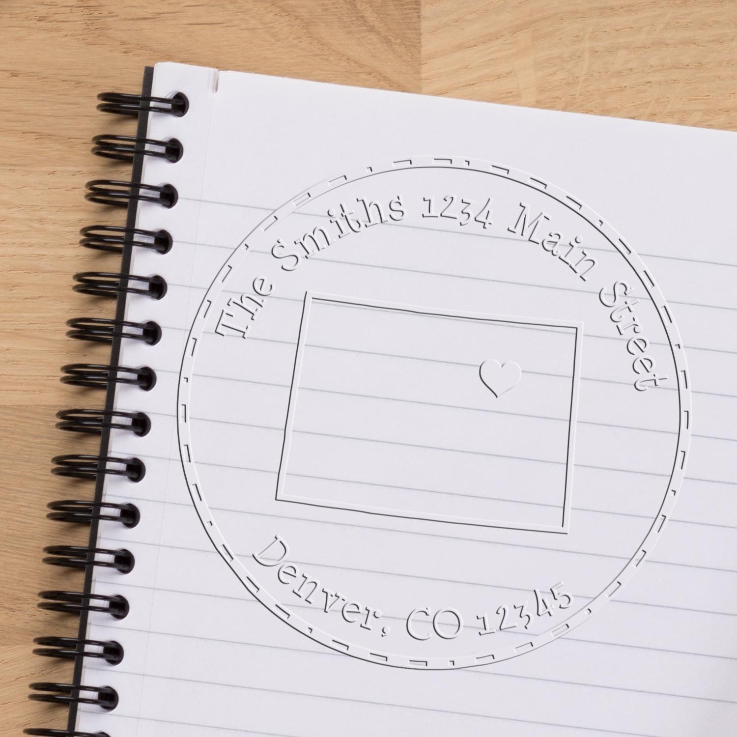 White spiral notebook featuring a circular address stamp seal with heart icon and black binding on a light wood desk.