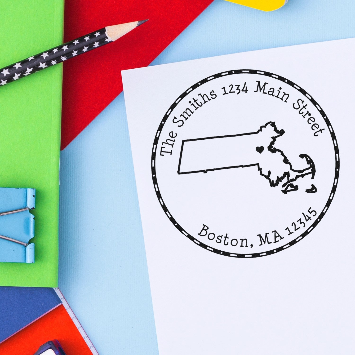 Custom black round address stamp and seal with Massachusetts outline on white paper, surrounded by bright green, red, blue, and yellow stationery.