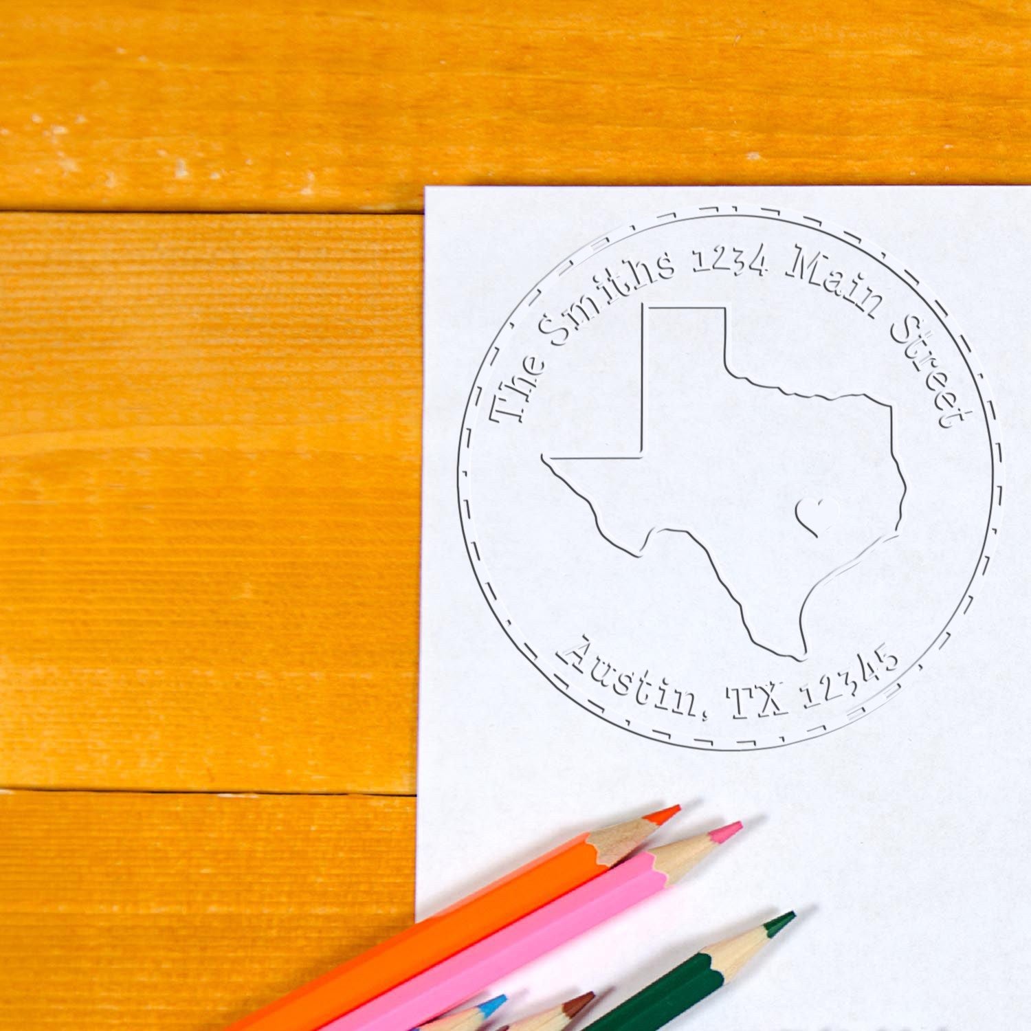 Custom stamp and seal with Texas outline and address, printed on white paper beside colorful pencils on a wood table.