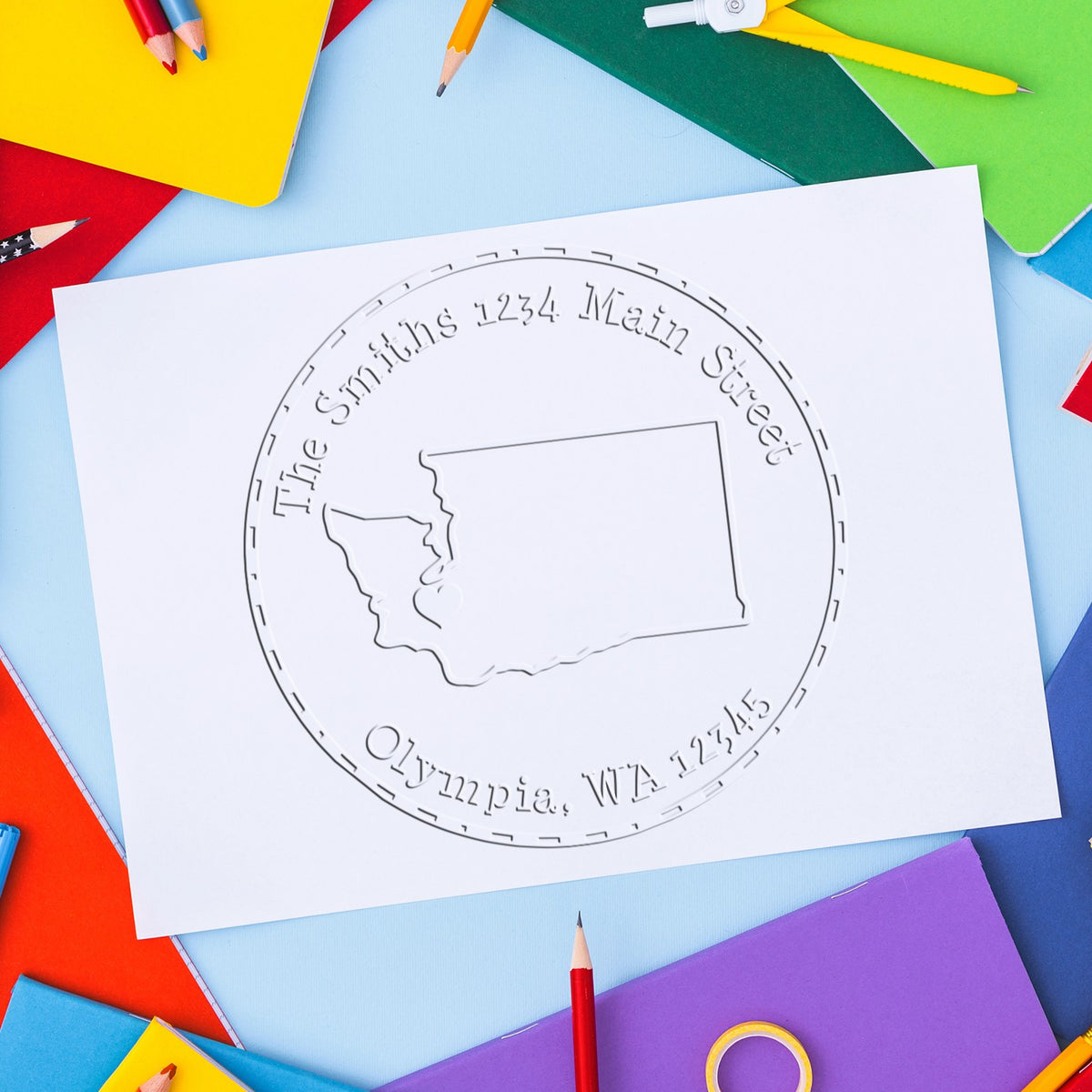 Embossed custom stamp and seal on white paper, with Washington state outline, surrounded by colorful notebooks and pencils.