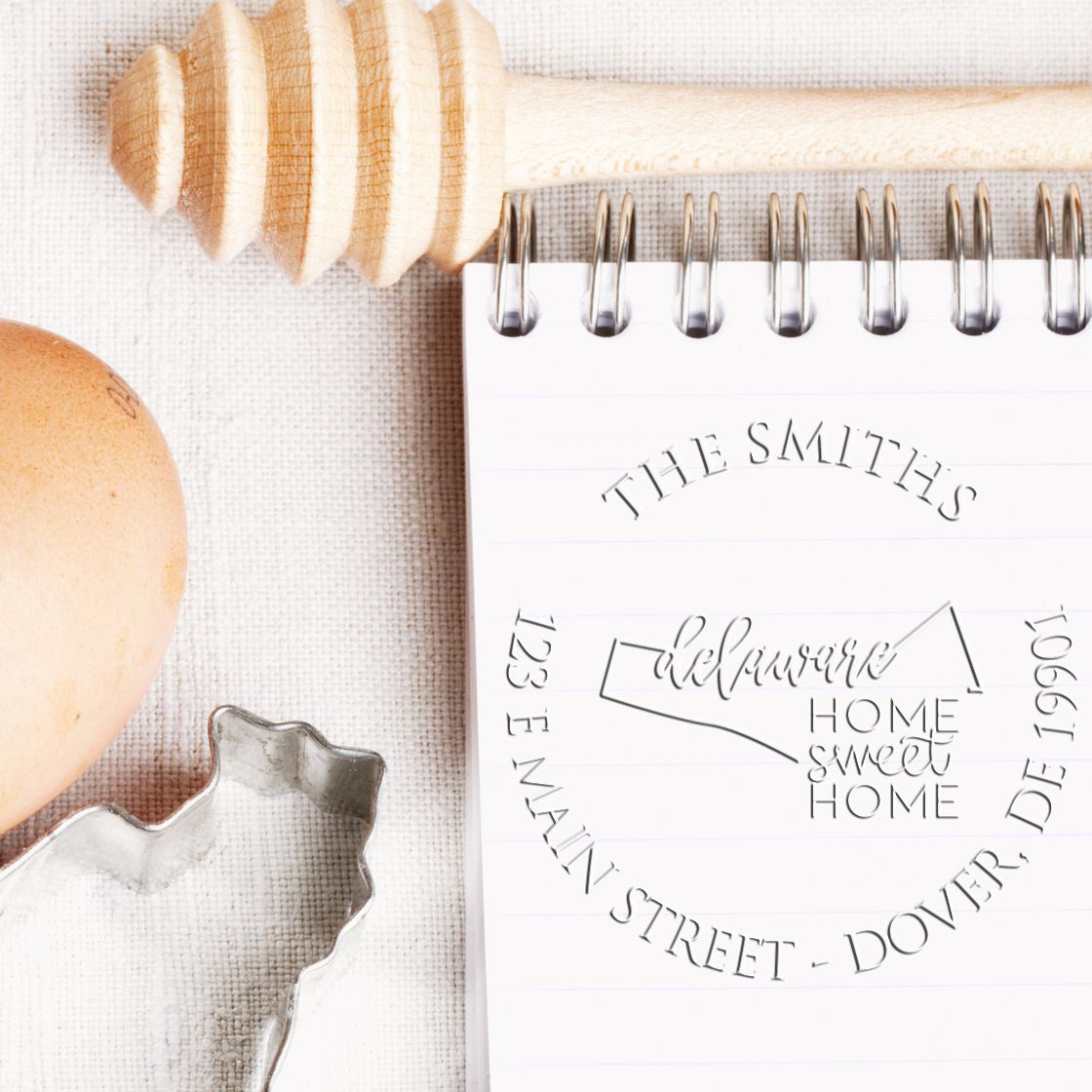Wooden stamp and metal seal cutter beside a spiral notebook with embossed The Smiths home sweet home on white linen.
