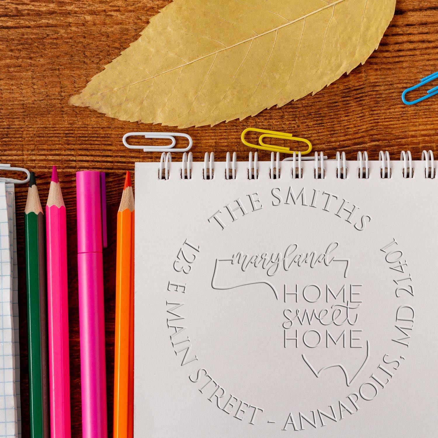 Custom embossed home seal on a white spiral notebook, with bright pencils, paper clips, and a tan leaf on a wood desk.