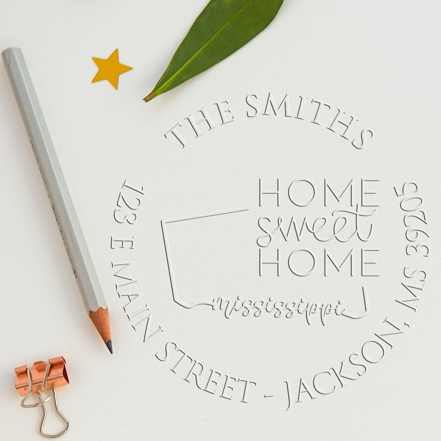 Custom stamp and embossing seal on white paper with silver text, green leaf, yellow star, pencil, and copper binder clip.