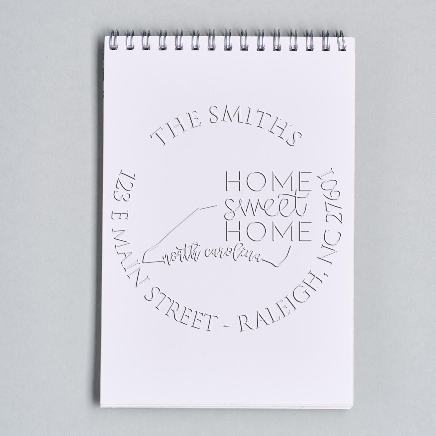 White spiral notepad with embossed stamp seal designs, including Home Sweet Home and The Smiths, on a light gray background.