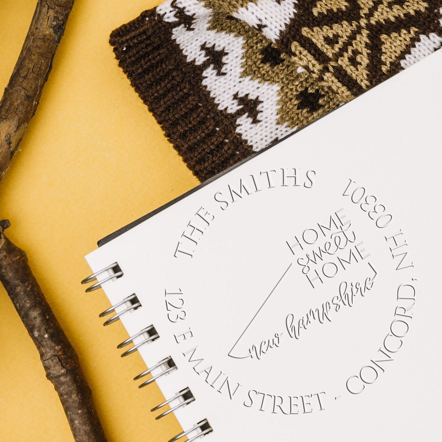 White spiral notebook with embossed custom stamp and seal designs, brown knit fabric, rustic branches, and a yellow background.