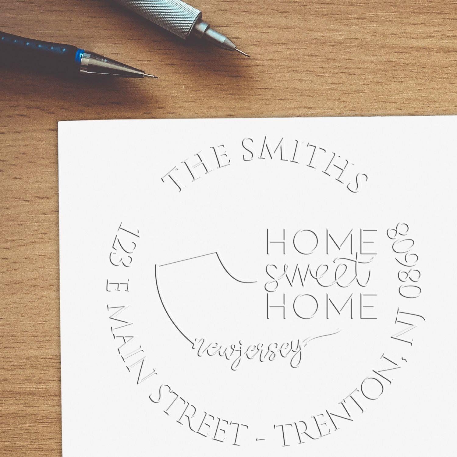 White custom stamp and seal proof on wood desk, with pens and embossed Home Sweet Home family crest and address.