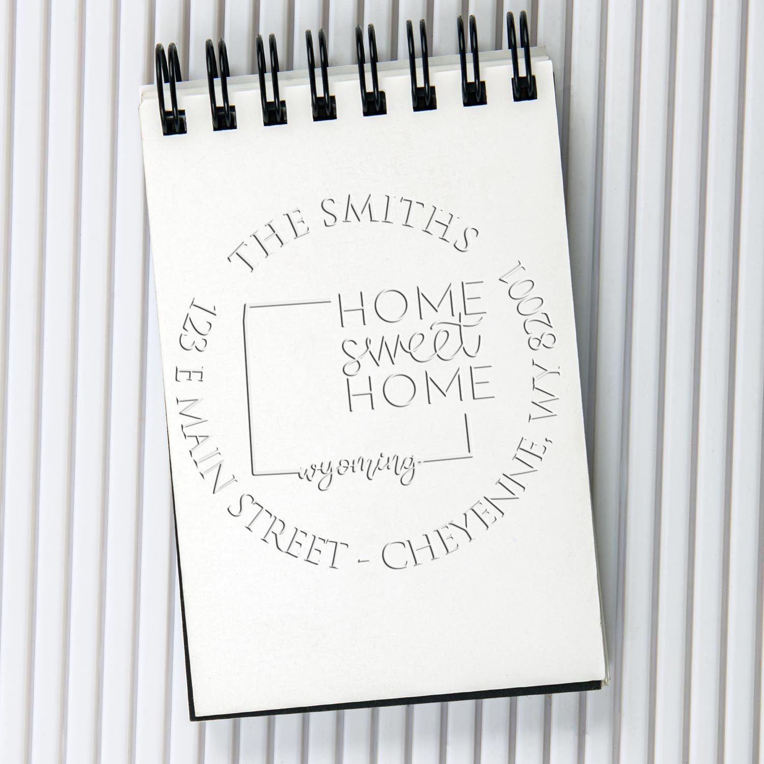 White spiral notebook displaying a custom address stamp seal design with embossed black text on a light gray background.