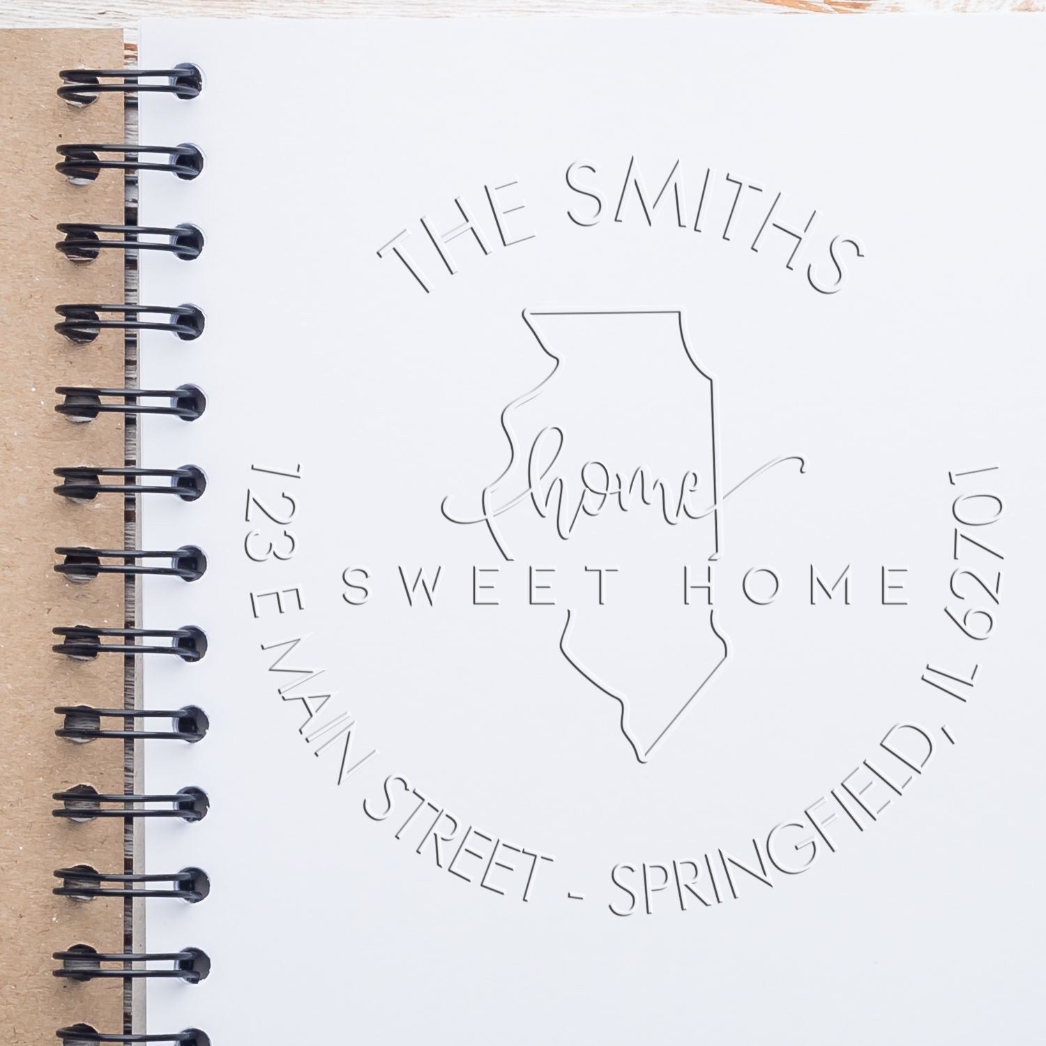 White embossed stamp seal on white spiral notebook with black binding, featuring The Smiths Sweet Home and Illinois address.