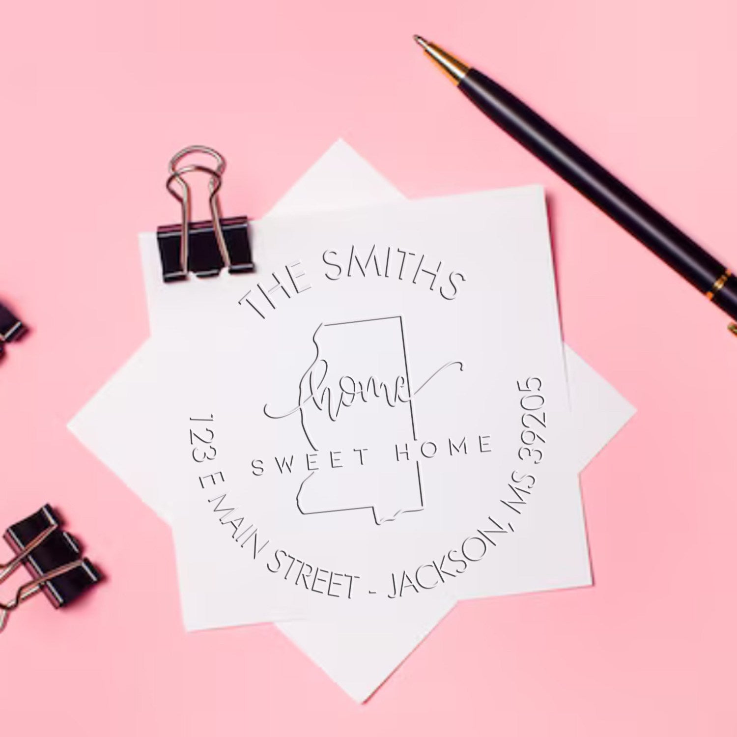 Custom seal stamp on white paper with pink background, black pen and binder clips, featuring The Smiths home address design.