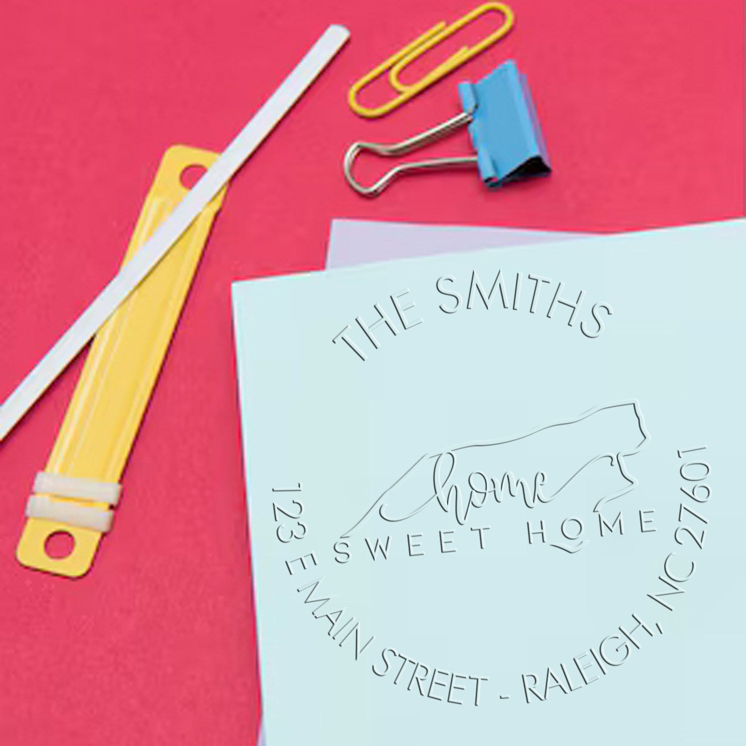 Custom embossed seal on light blue paper reading Home Sweet Home, with gold paperclips, blue binder clip, and red background.