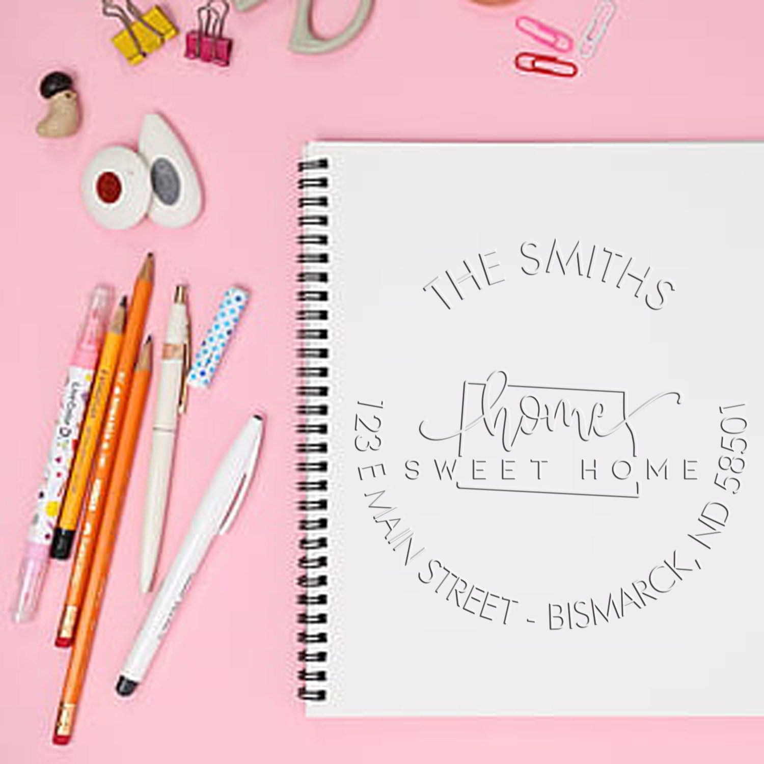 Personalized embossed address seal on a white spiral notebook, with pink background, pens, and binder clips.