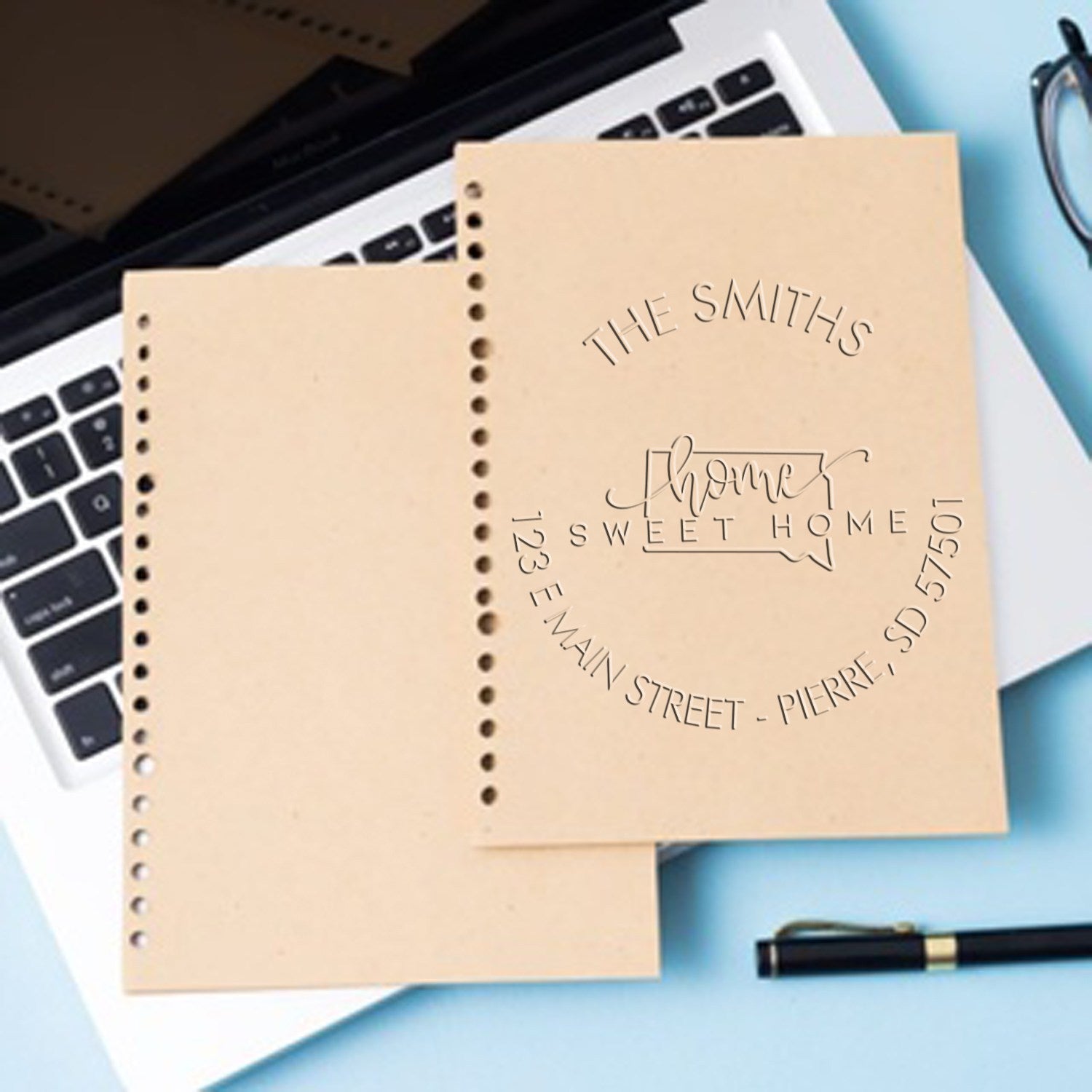 Beige custom stamp and seal samples with embossed family name and address on perforated sheets over a laptop on a blue desk.