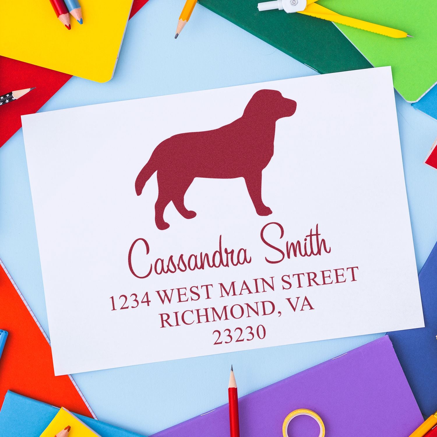 Burgundy custom stamp and seal with dog silhouette on white card, surrounded by colorful notebooks and pencils.