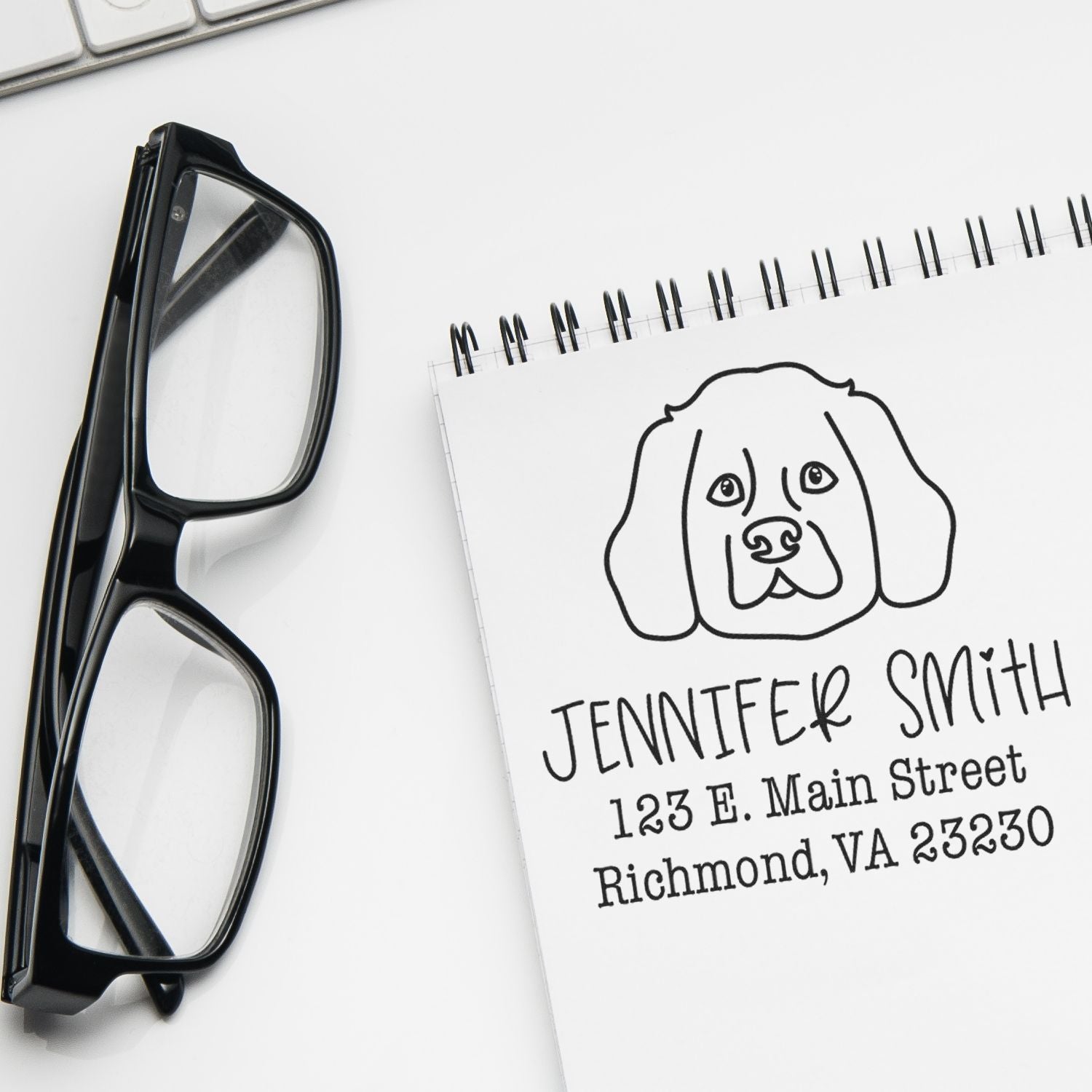 Black eyeglasses beside a personalized dog stamp and seal notepad with name and address on a white desk.