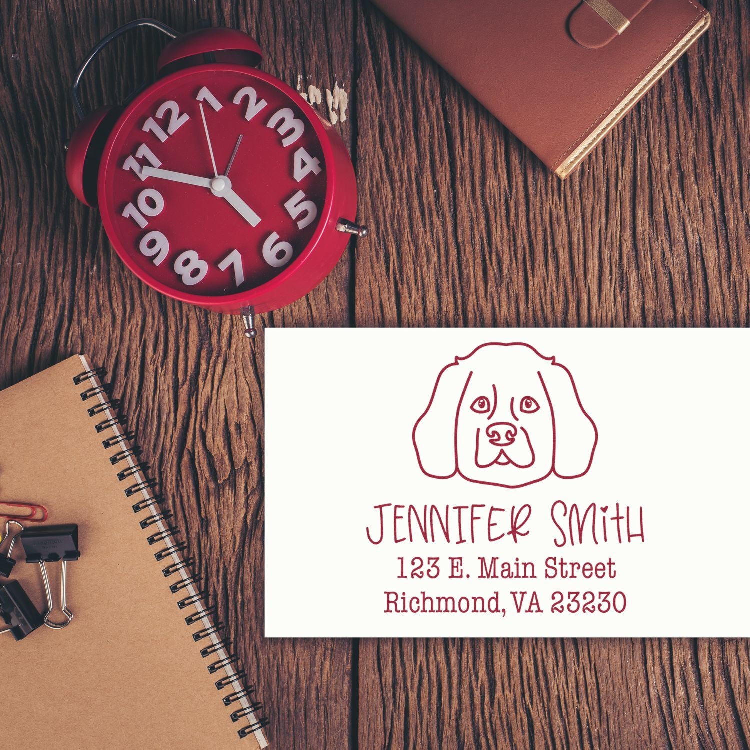 Maroon custom address stamp and seal with dog logo and Jennifer Smith text on white card, beside a red clock and brown notebook.
