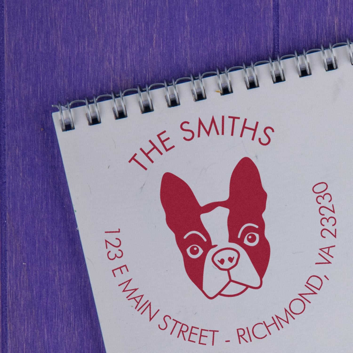 Custom business stamp and seal on white spiral notebook, red dog logo and address details against a purple background.