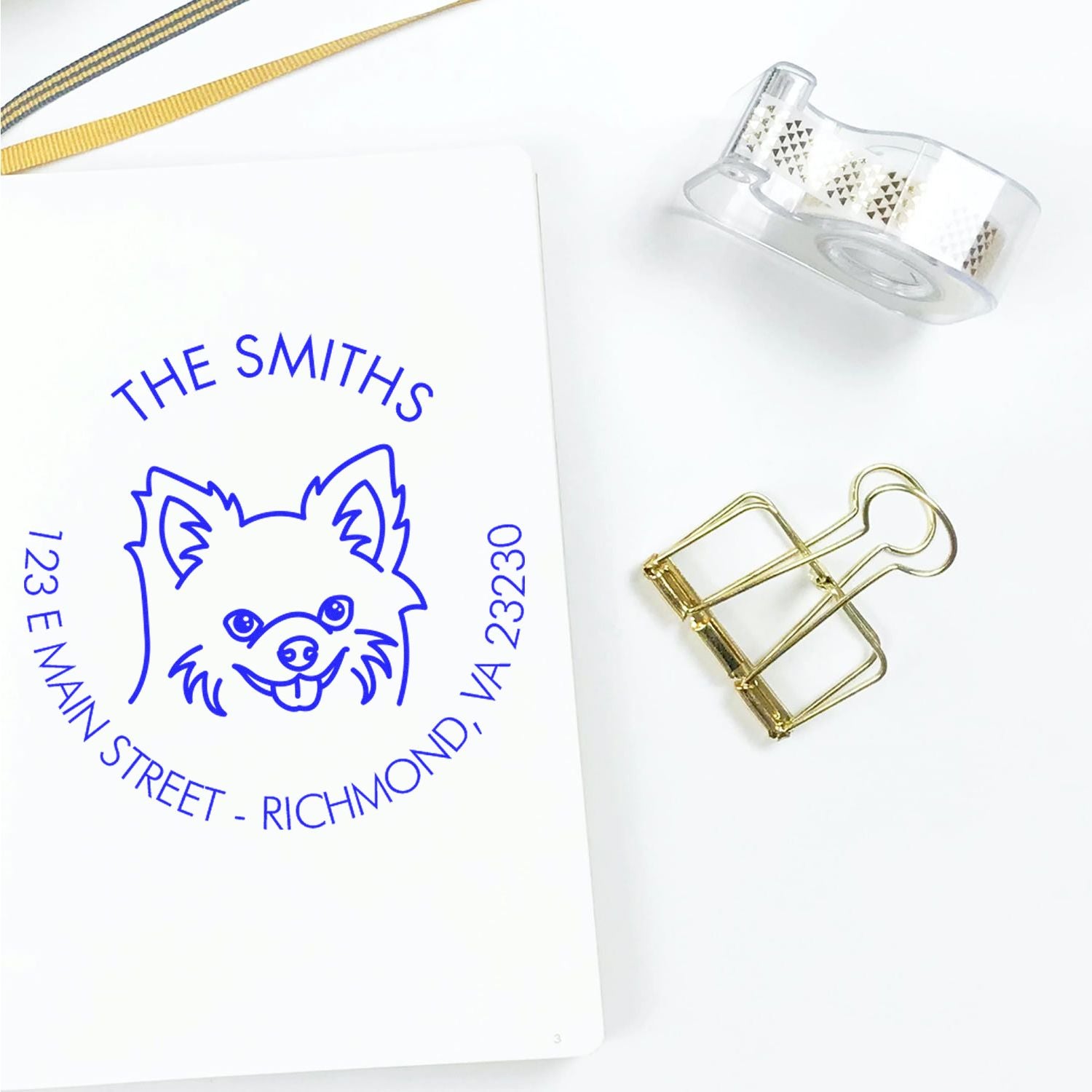 Blue custom dog address stamp on white paper, with clear seal tape roll, gold clip, and gold ribbon accents.